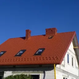 Dom w trakcie budowy z nowym, czerwonym dachem z dachówką, trzema oknami dachowymi i dwoma kominami z cegły, na tle błękitnego nieba.