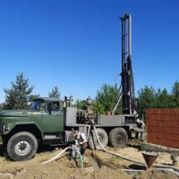 Wiertnica geologiczna na podwoziu ciężarówki ZIL-131, pracownik obsługujący pompę wodną w wykopie, w tle mur z czerwonej cegły i drzewa.