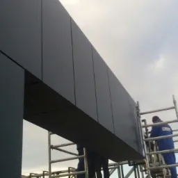 Montaż paneli elewacyjnych w kolorze grafitowym, widoczni pracownicy na rusztowaniu, pochmurne niebo w tle.