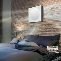 Nowoczesny, kwadratowy oczyszczacz powietrza z podświetleniem LED zamontowany na ceglanej ścianie w sypialni, widoczne promienie światła słonecznego wpadające przez okno z drewnianymi okiennicami.