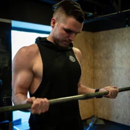 Mężczyzna w czarnej koszulce bez rękawów z kapturem, ćwiczący biceps z długim gryfem w siłowni, widoczne napięte mięśnie ramion.