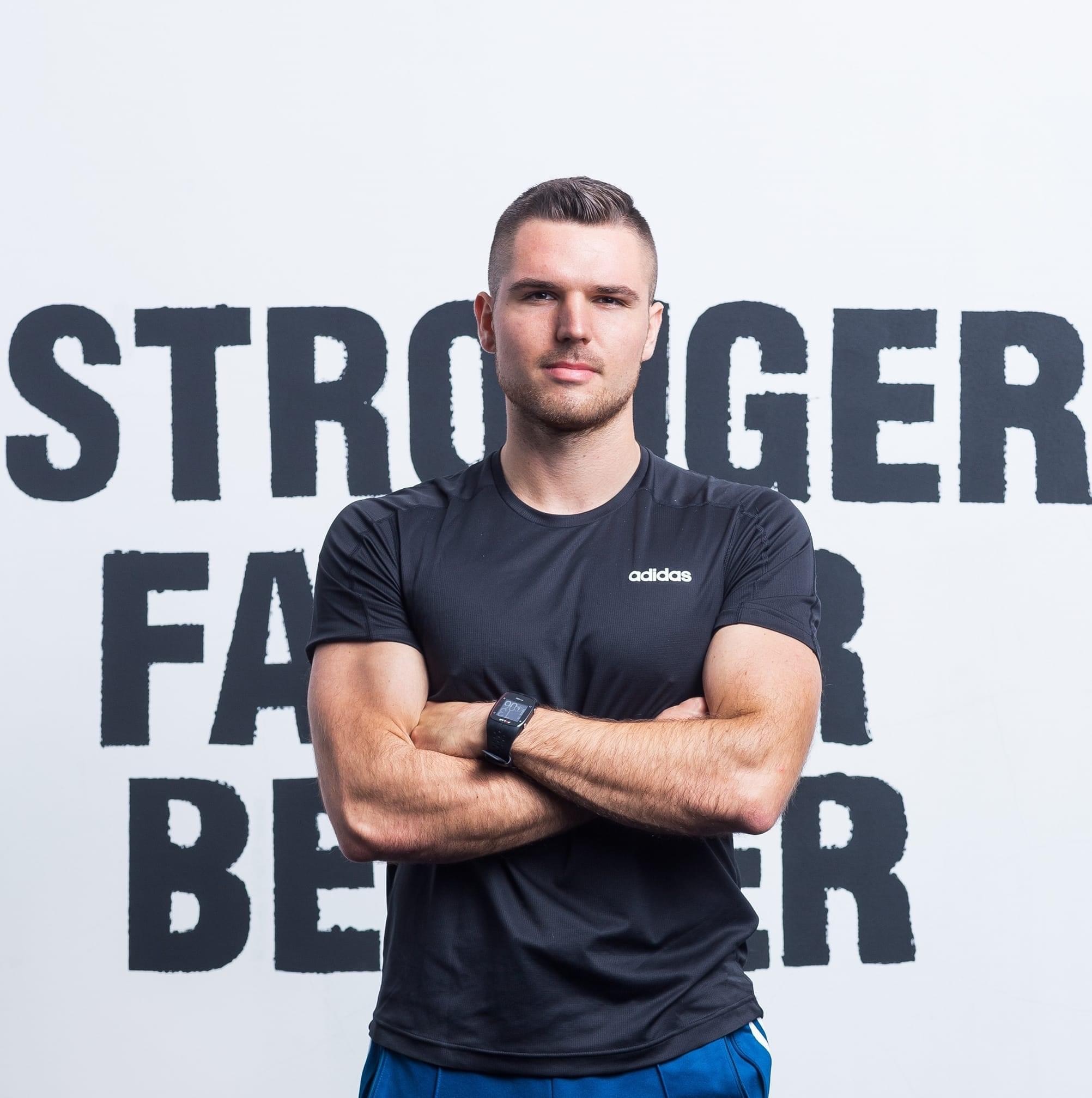 Młody mężczyzna w sportowym stroju Adidas z założonymi rękami na tle ściany z napisem 'STRONGER FASTER BETTER', widoczny zegarek sportowy.