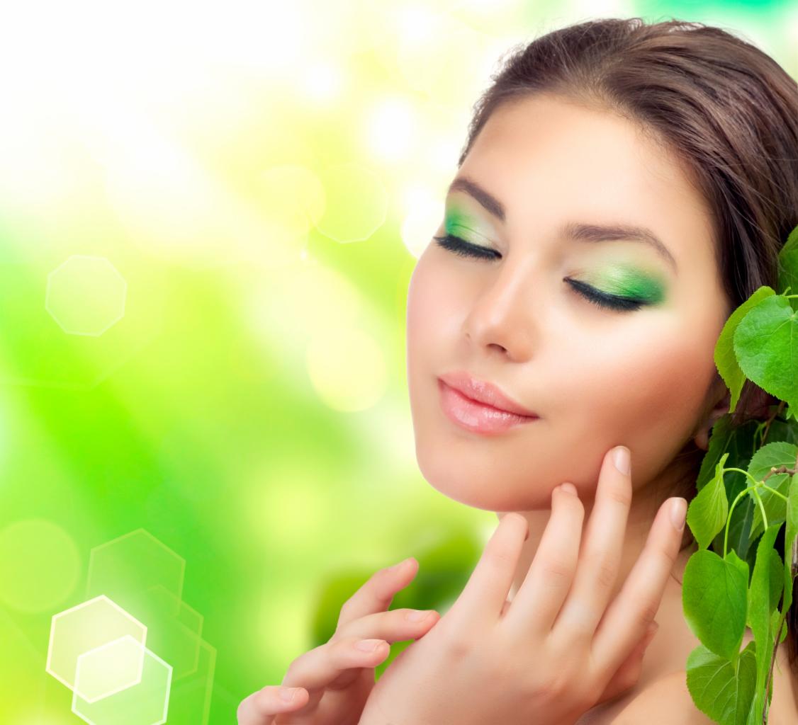 Portret kobiety z zamkniętymi oczami, w zielonym makijażu oczu, z gałązką zielonych liści przy twarzy, na rozmytym zielono-żółtym tle.