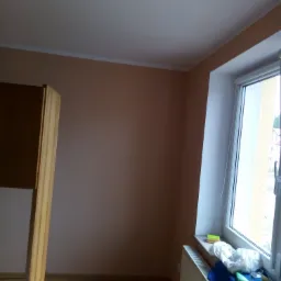 Pokój z beżowymi ścianami przed remontem, widoczne okno, grzejnik i fragment mebla.