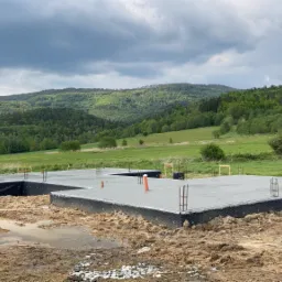 Wylana płyta fundamentowa na tle zielonych wzgórz, widoczne wystające pręty zbrojeniowe i rury kanalizacyjne, teren wokół w stanie surowym.