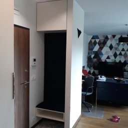 Zabudowa Wnętrz Artur Retke - Przedpokój w Chotomowie z zabudowaną szafą wnękową z siedziskiem i półką, obok drewniane drzwi wejściowe, w tle widoczny salon z geometryczną tapetą.