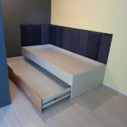 Wysuwane łóżko z szufladą i wezgłowiem z ciemnej tapicerki panelowej na tle ściany w dwóch odcieniach.