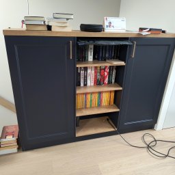 Zabudowa Wnętrz Artur Retke - Niebieska szafka z drewnianym blatem i półkami wypełnionymi książkami, dodatkowo książki ułożone na blacie i pod szafką, widoczny kabel zasilający na podłodze.