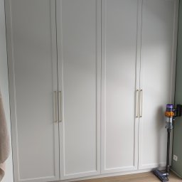 Zabudowa Wnętrz Artur Retke - Czterodrzwiowa szafa wnękowa w odcieniach szarości z podłużnymi złotymi uchwytami, na podłodze panele, obok odkurzacz pionowy.