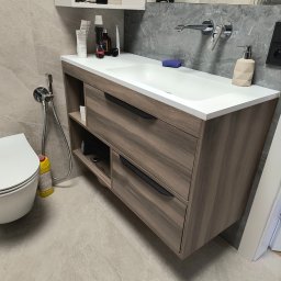 Zabudowa Wnętrz Artur Retke - Nowoczesna łazienka z szafką pod umywalkę w odcieniach drewna, białą umywalką i baterią ścienną. Widoczne kosmetyki i akcesoria łazienkowe na blacie. Obok toaleta i bidetka.