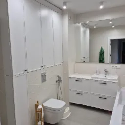 Nowoczesna łazienka z zabudową meblową w bieli, szafką pod umywalką z szufladami i wysoką szafą. Minimalistyczny design, szare płytki podłogowe.