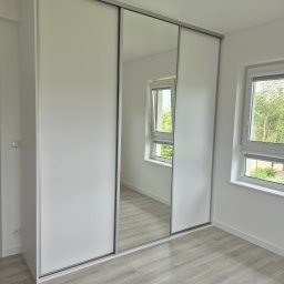 Zabudowa Wnętrz Artur Retke - Biała szafa wnękowa z przesuwnymi drzwiami i lustrem, odbijająca okno z widokiem na zieleń, na tle jasnej podłogi w nowym wnętrzu.