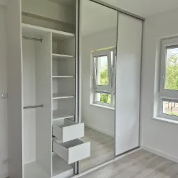 Biała szafa przesuwna z lustrem, otwarte półki i szuflady, jasne wnętrze, widok na okno z zielenią w tle, minimalistyczny design.