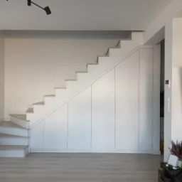 Białe schody z wbudowaną szafą w minimalistycznym wnętrzu. Nowoczesne oświetlenie sufitowe i jasna podłoga. Widok na telewizor i dekoracje.