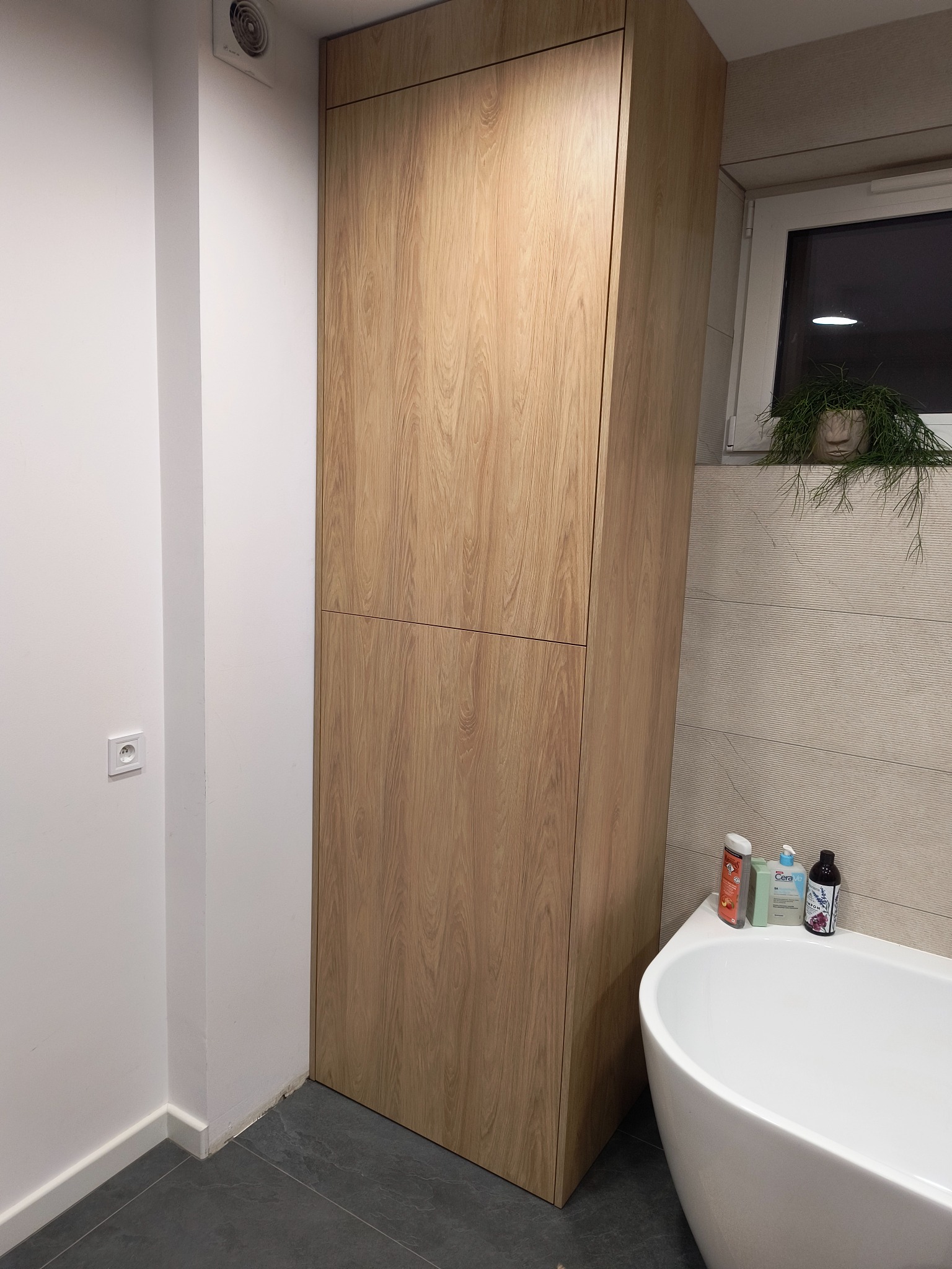 Wysoka, dwudrzwiowa szafa z jasnego drewna w łazience, stojąca obok wanny i okna z rośliną doniczkową, na szarych płytkach podłogowych.