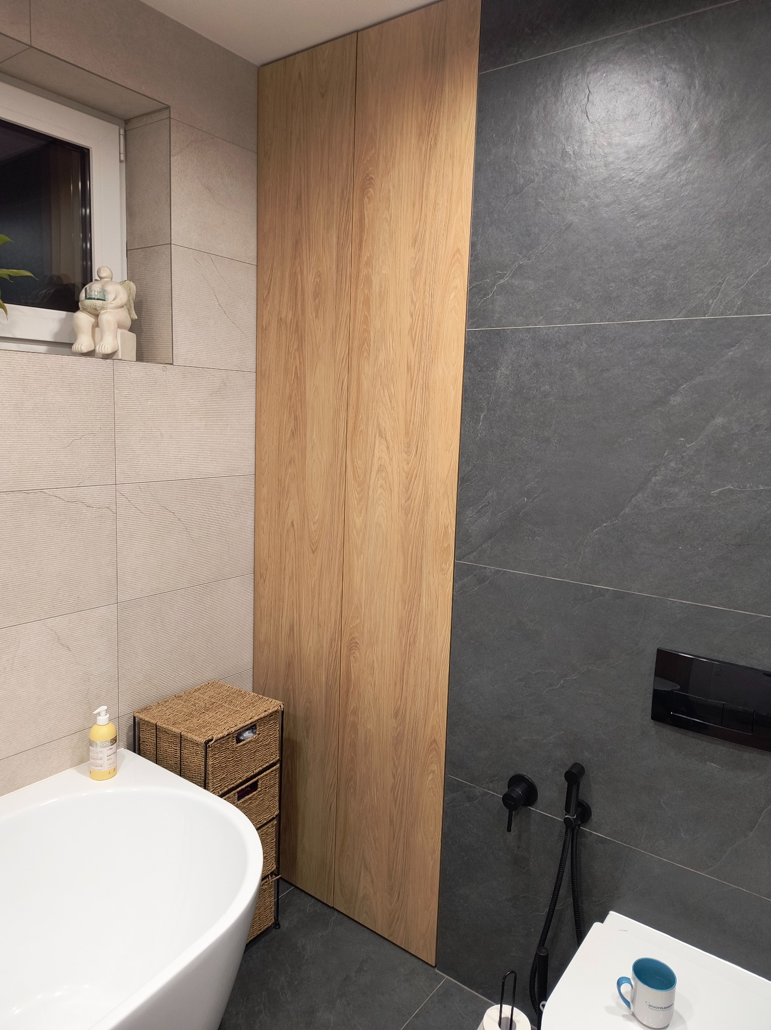 Wysoka, drewniana szafa w łazience z szarymi płytkami, obok wanna i wiklinowa komoda z szufladami, na parapecie figurka anioła.