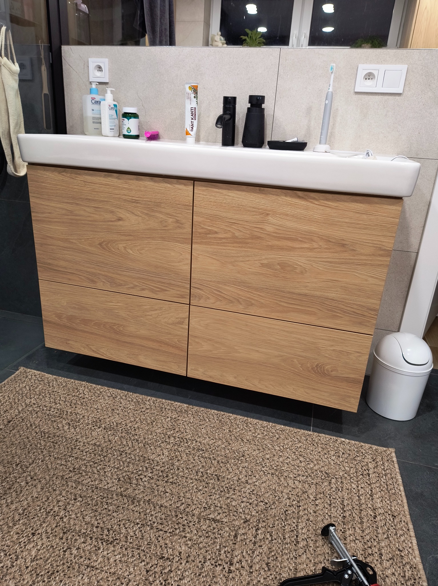 Łazienka z drewnianą szafką pod umywalką, na blacie kosmetyki i szczoteczka elektryczna, na podłodze dywanik i narzędzie do montażu