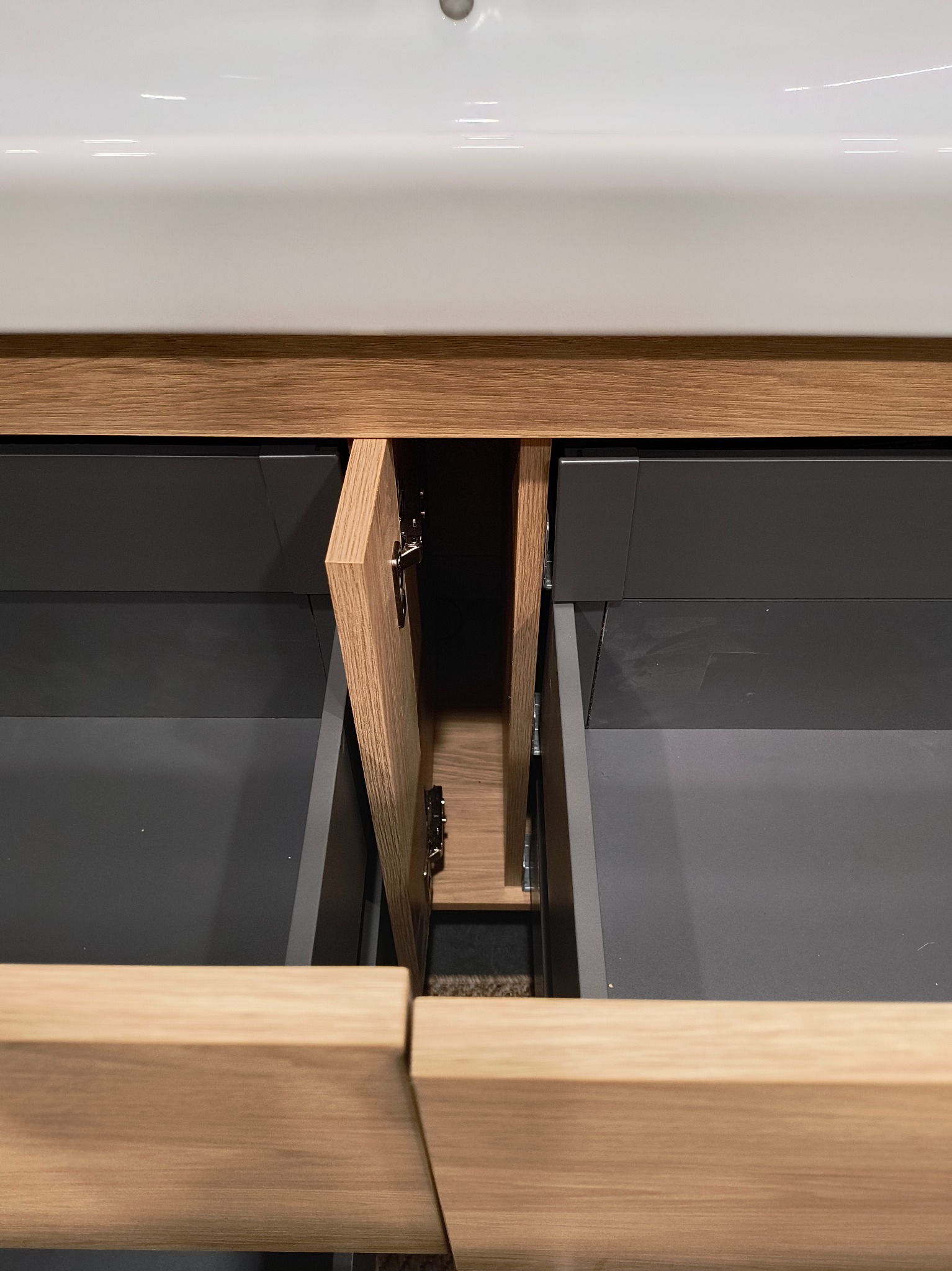 Otwarta szafka pod umywalką z drewnianymi frontami i szarymi szufladami, widoczne zawiasy.