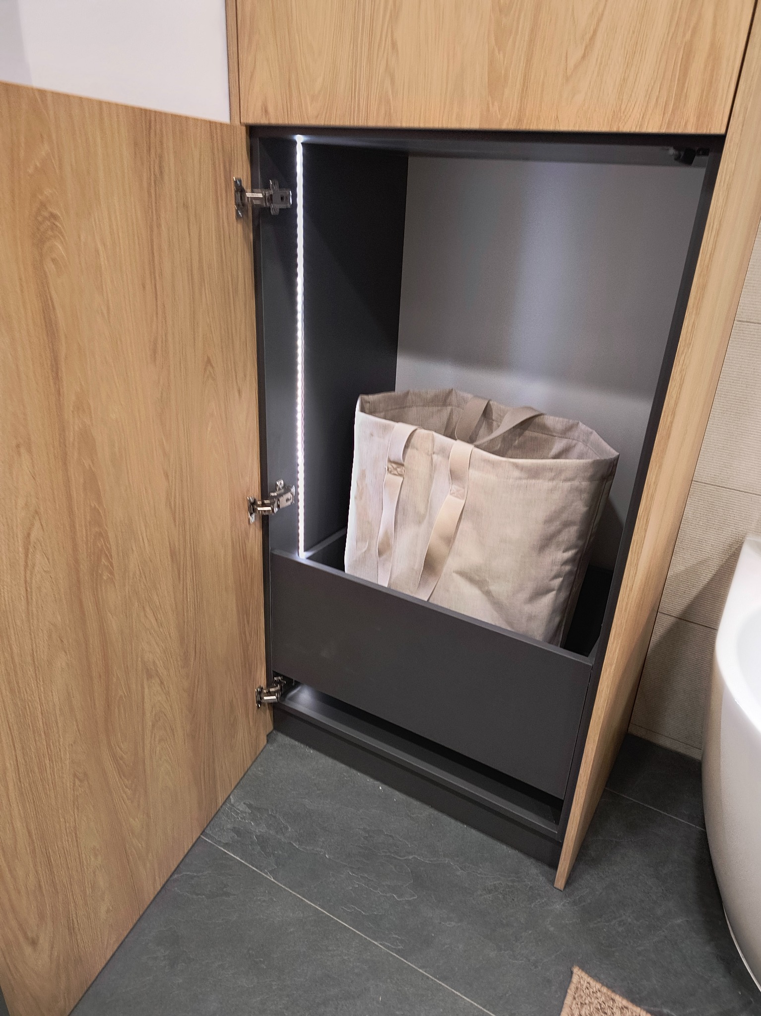 Otwarta szafka z drewnopodobnymi frontami, podświetlona listwą LED, z wysuwaną szufladą zawierającą beżową torbę na zakupy.