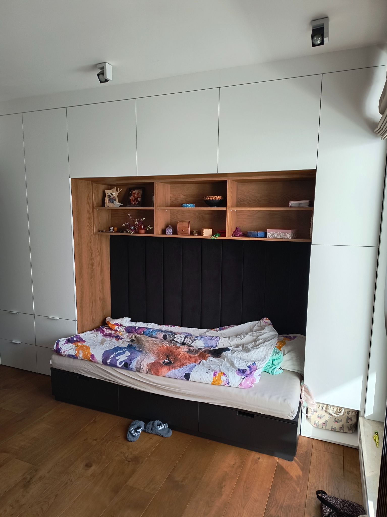 Biała szafa wnękowa z drewnianymi półkami nad łóżkiem z czarnym zagłówkiem i kolorową pościelą. Na podłodze leżą kapcie.