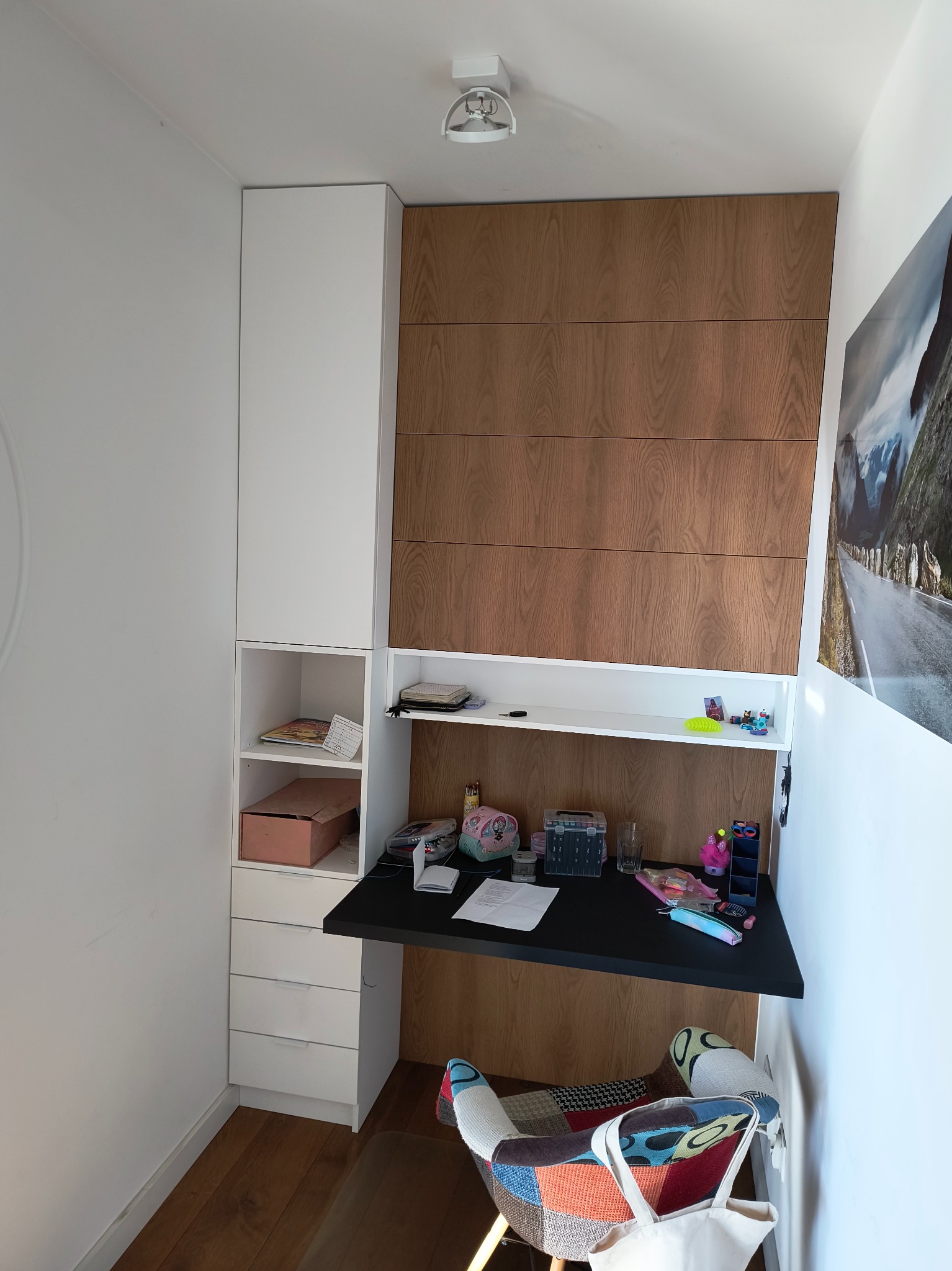 Zintegrowane biurko z szafkami wnękowymi, blat w kolorze czarnym, fronty szafek w odcieniach drewna, krzesło patchworkowe w tle, oświetlenie sufitowe.