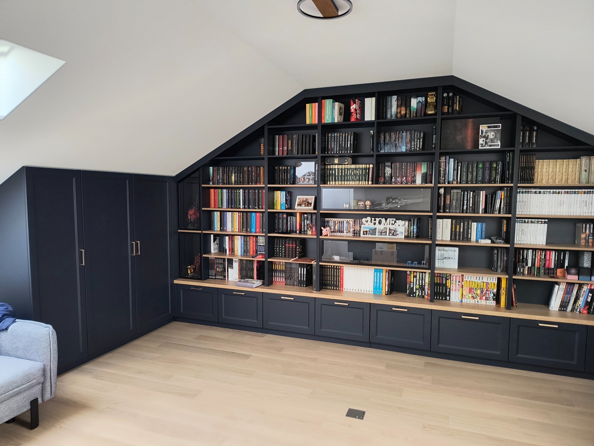 Czarna zabudowa meblowa pod skosem dachu z szafami i regałami na książki, jasna drewniana podłoga, widoczny fragment jasnoszarego fotela.