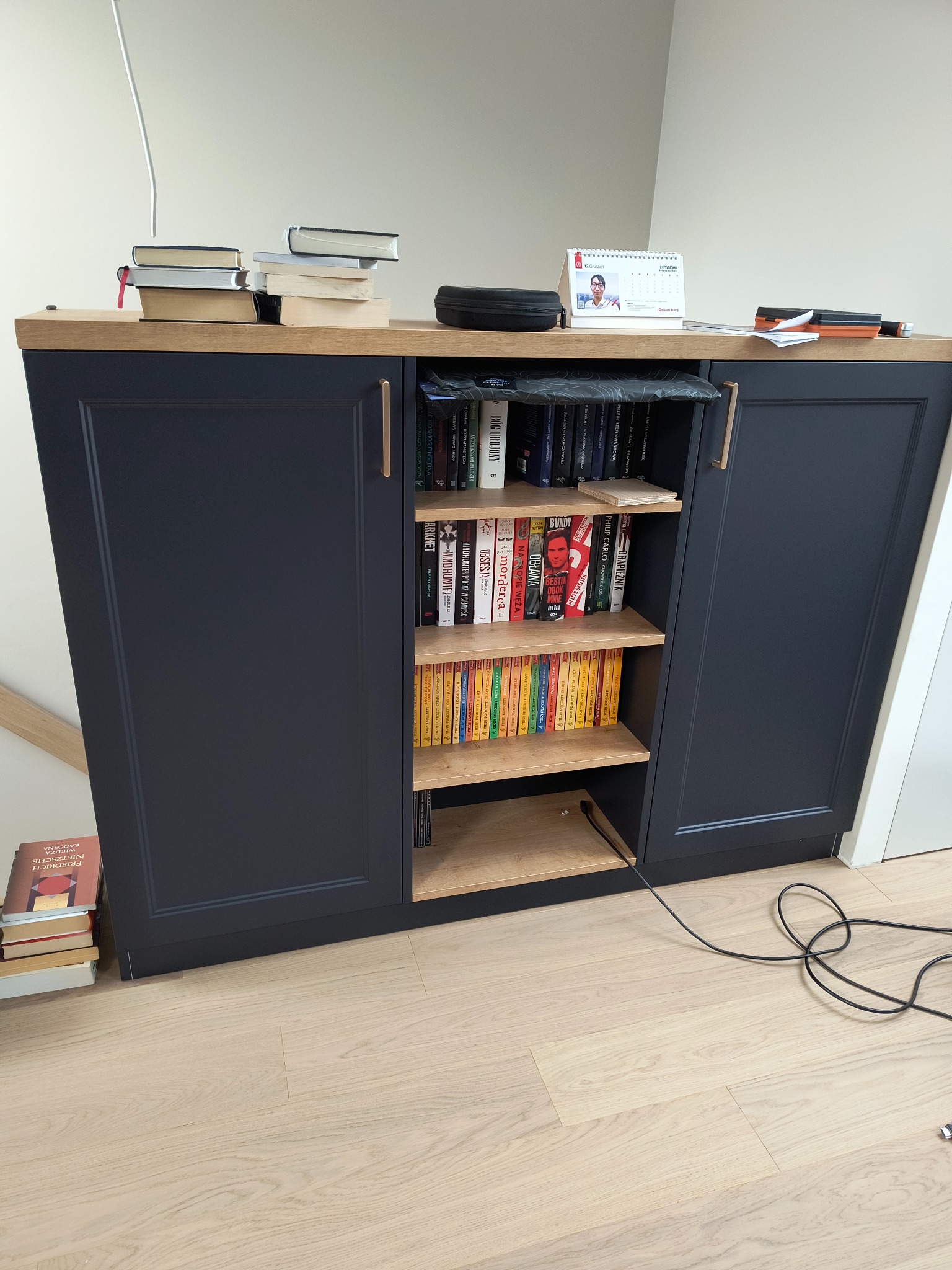 Niebieska szafka z drewnianym blatem i półkami wypełnionymi książkami, dodatkowo książki ułożone na blacie i pod szafką, widoczny kabel zasilający na podłodze.