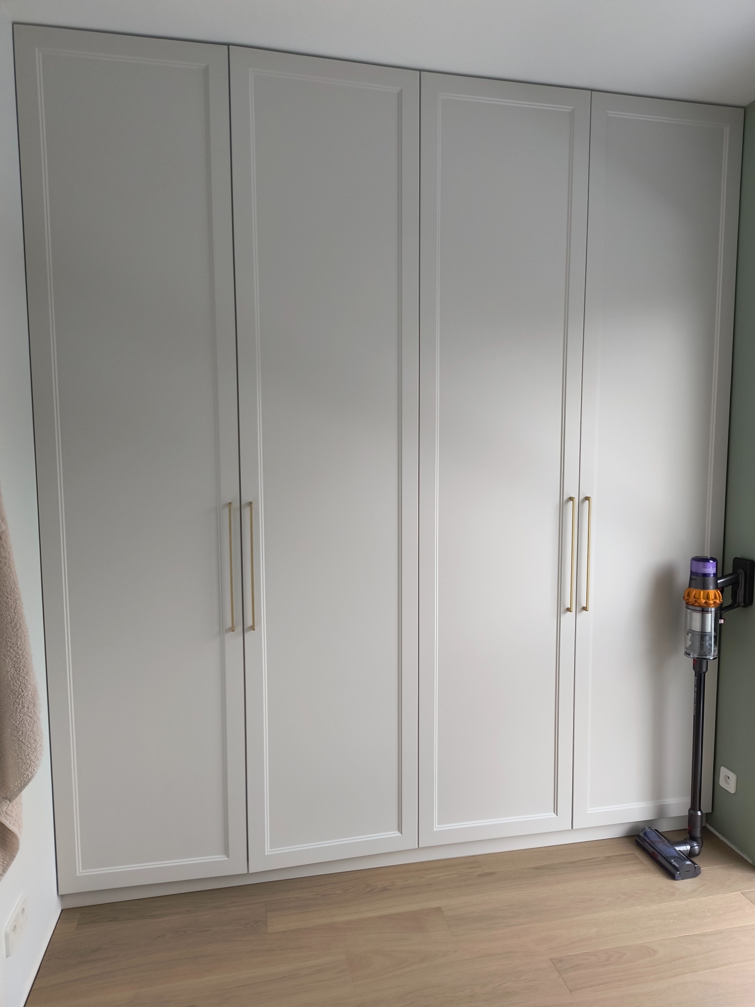Czterodrzwiowa szafa wnękowa w odcieniach szarości z podłużnymi złotymi uchwytami, na podłodze panele, obok odkurzacz pionowy.