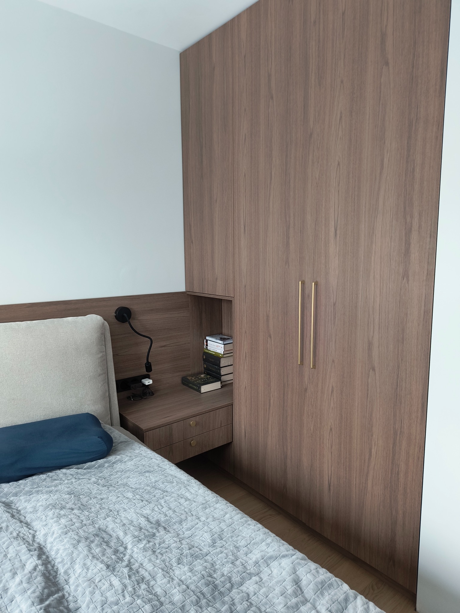 Drewniana szafa wnękowa zintegrowana z szafką nocną i półkami, widok z perspektywy łóżka z niebieską poduszką i szarą narzutą.