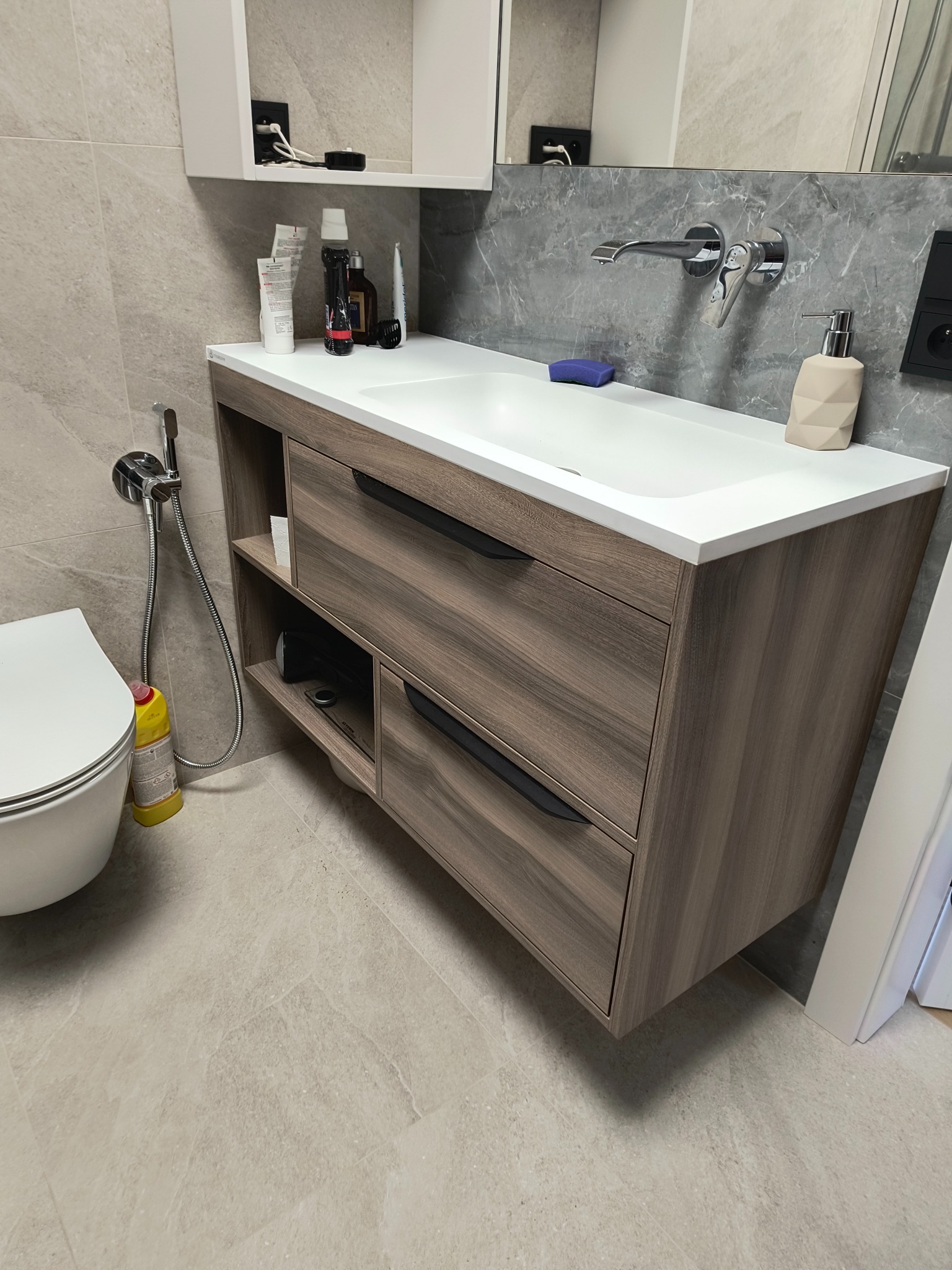 Nowoczesna łazienka z szafką pod umywalkę w odcieniach drewna, białą umywalką i baterią ścienną. Widoczne kosmetyki i akcesoria łazienkowe na blacie. Obok toaleta i bidetka.