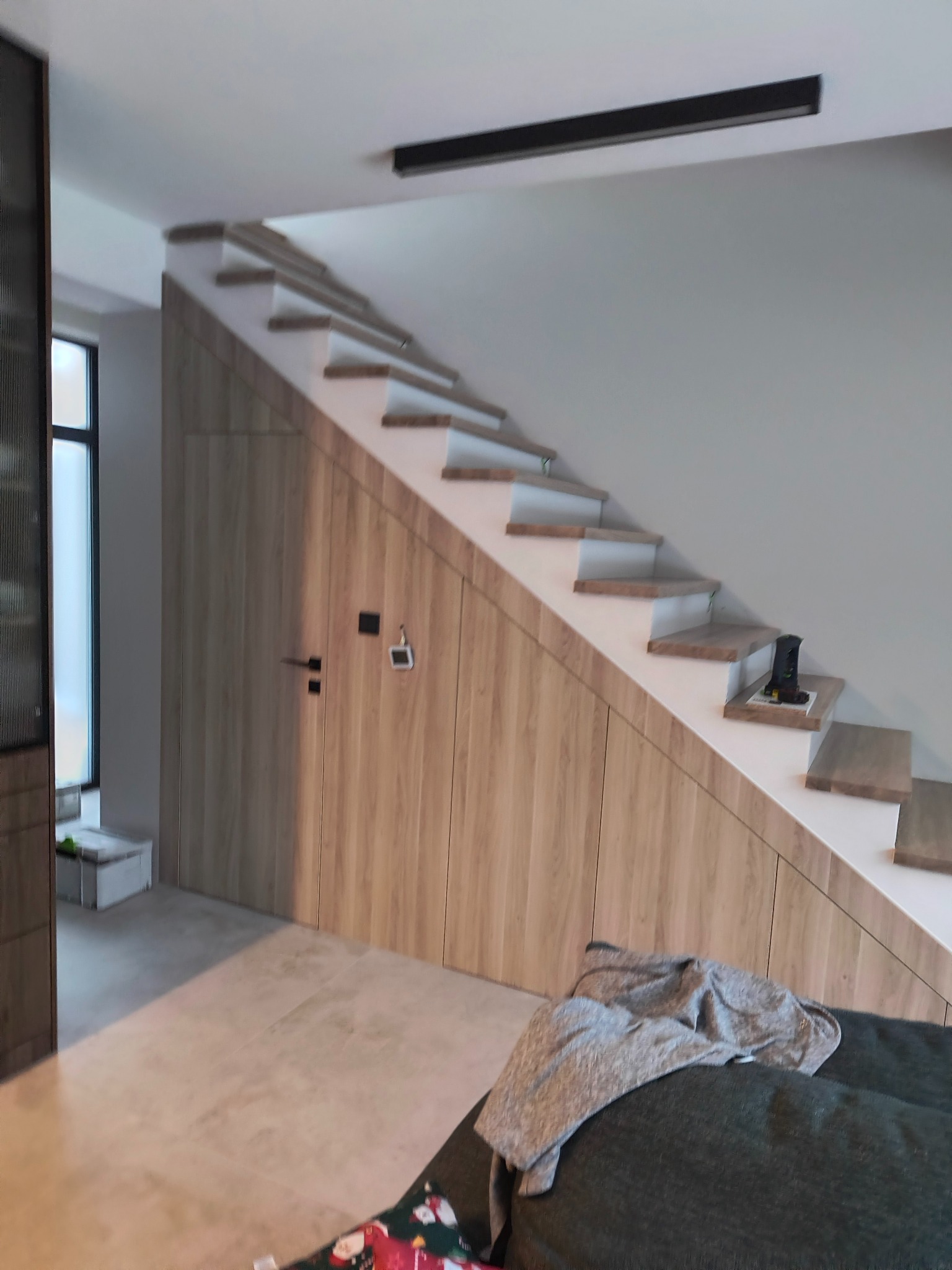 Drewniane schody ze schowkiem pod spodem, zrealizowane w Chotomowie, z widocznym oświetleniem sufitowym i fragmentem umeblowania w dolnej części kadru.