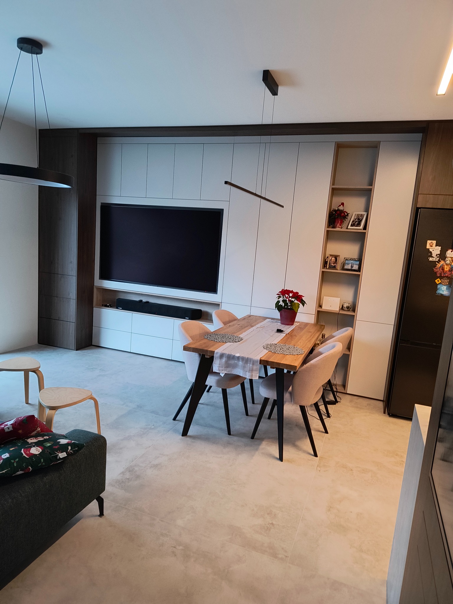 Nowoczesna jadalnia z drewnianym stołem, krzesłami tapicerowanymi w kolorze beżowym i zabudową meblową z białymi frontami i elementami w kolorze ciemnego drewna, w tle telewizor i półki z dekoracjami.