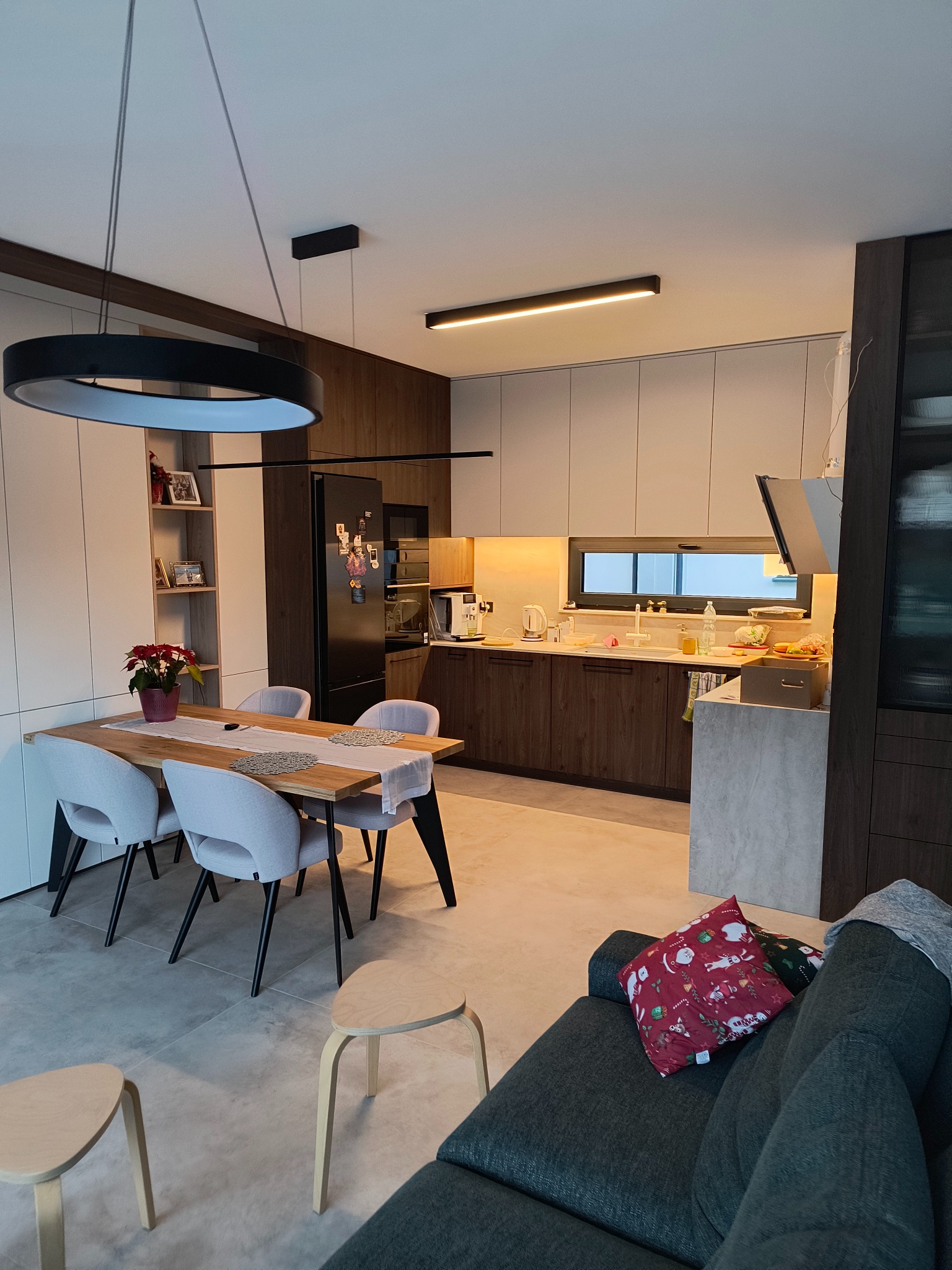 Nowoczesna kuchnia z drewnianymi i białymi frontami, oświetleniem LED i stołem jadalnym z szarymi krzesłami w tle sofy z dekoracyjnymi poduszkami.