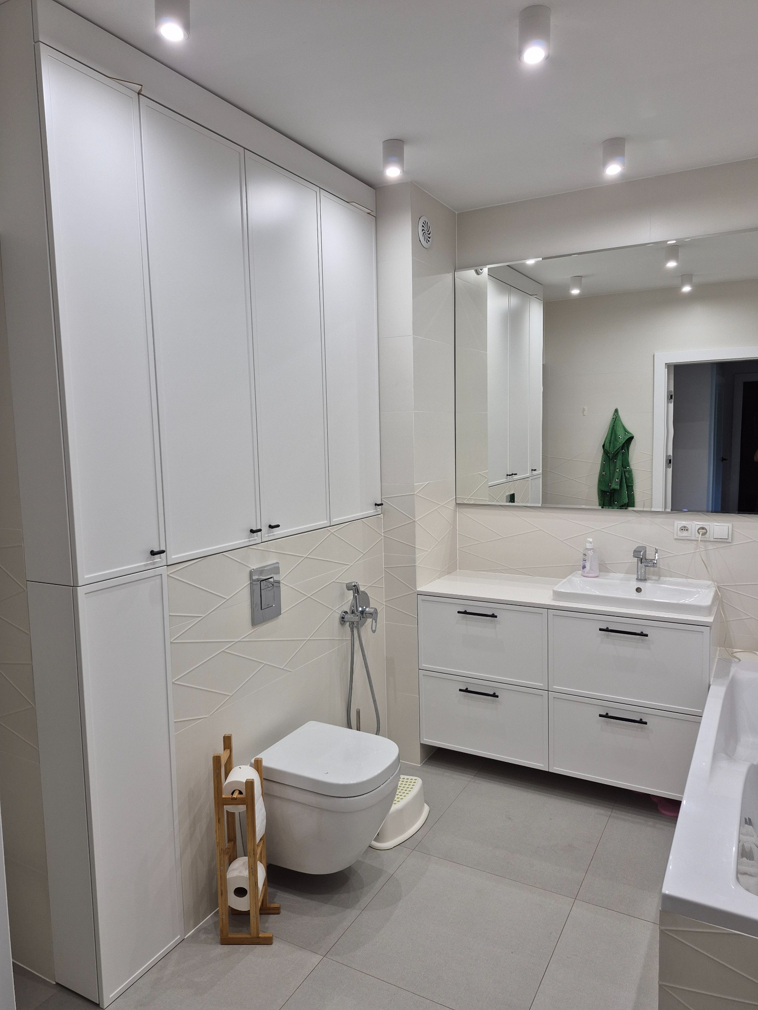 Nowoczesna łazienka z zabudową meblową w bieli, szafką pod umywalką z szufladami i wysoką szafą. Minimalistyczny design, szare płytki podłogowe.