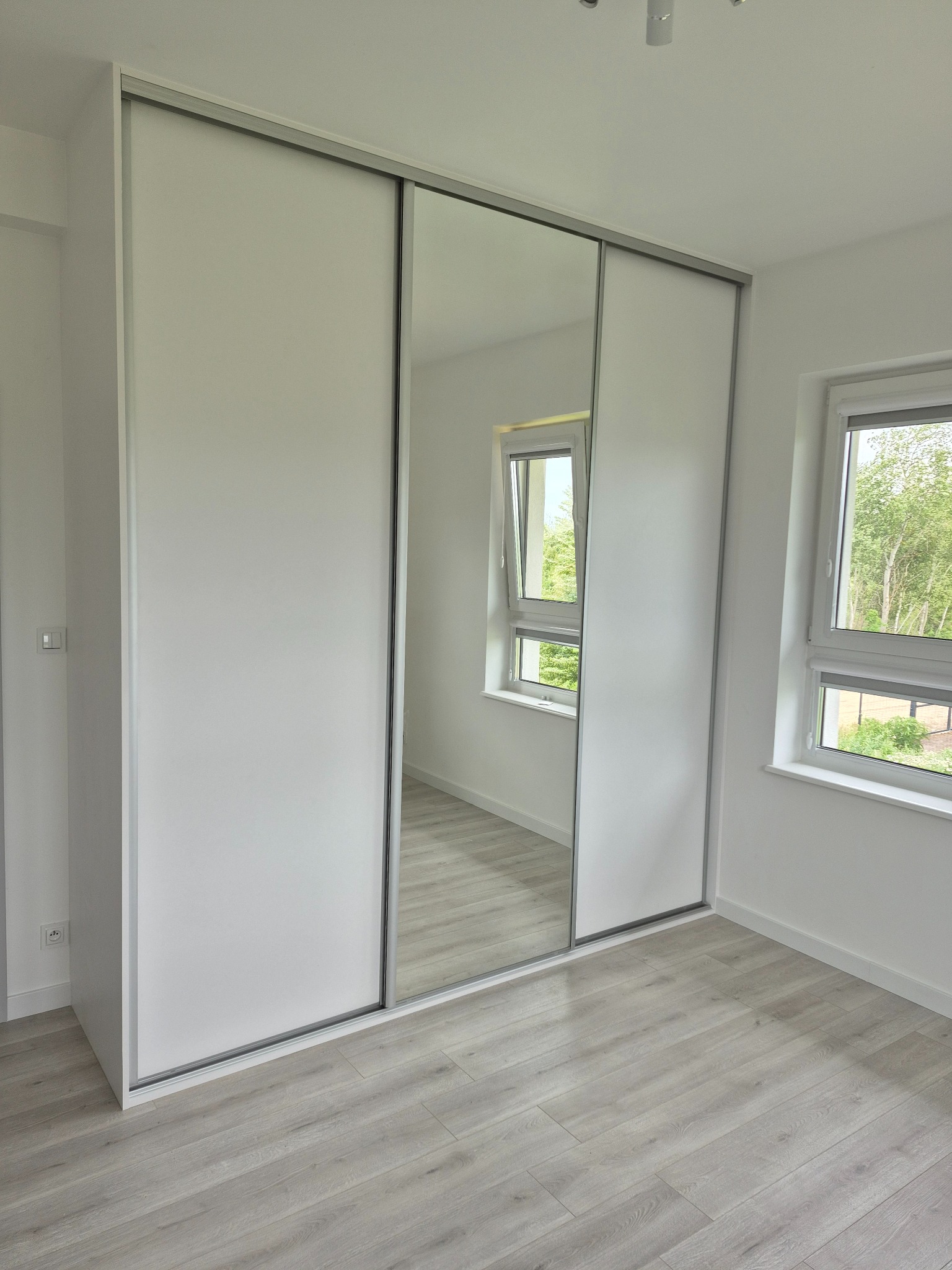 Biała szafa wnękowa z przesuwnymi drzwiami i lustrem, odbijająca okno z widokiem na zieleń, na tle jasnej podłogi w nowym wnętrzu.