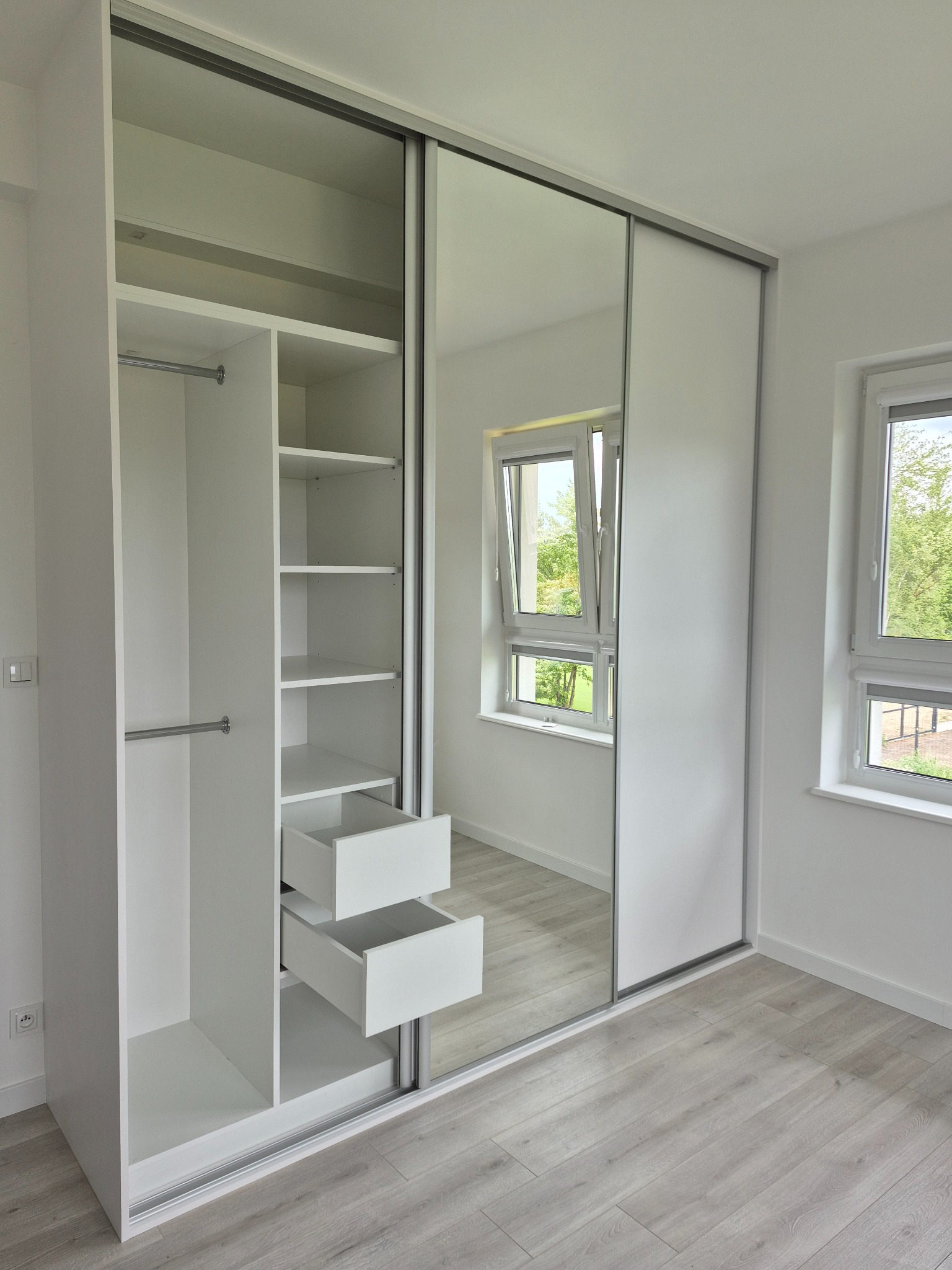 Biała szafa przesuwna z lustrem, otwarte półki i szuflady, jasne wnętrze, widok na okno z zielenią w tle, minimalistyczny design.