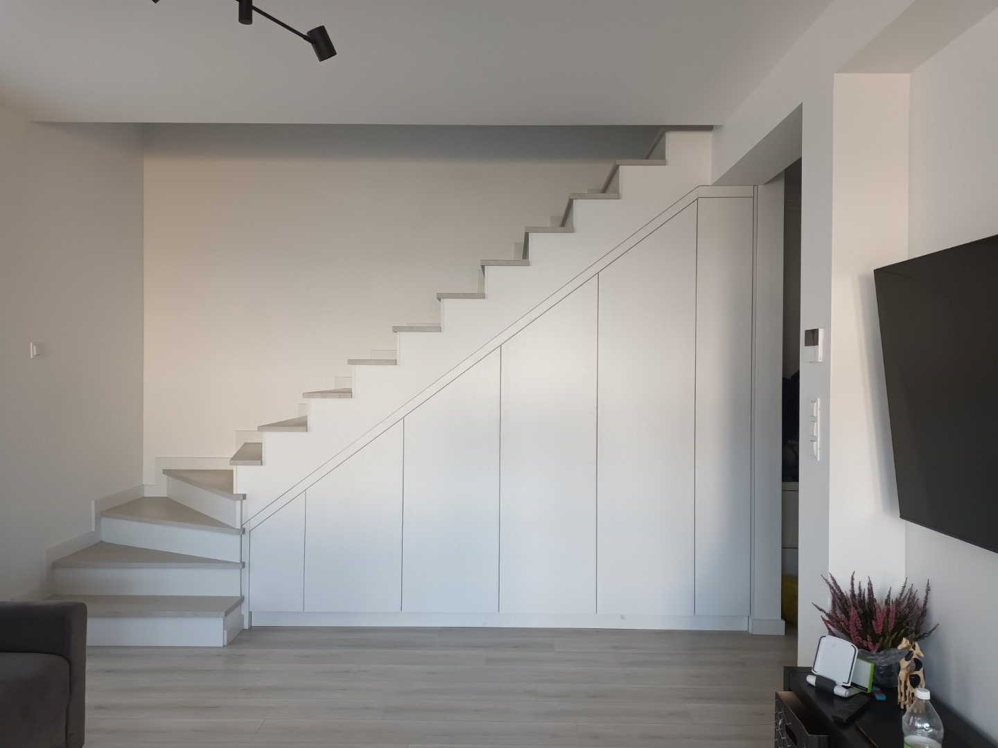 Białe schody z wbudowaną szafą w minimalistycznym wnętrzu. Nowoczesne oświetlenie sufitowe i jasna podłoga. Widok na telewizor i dekoracje.