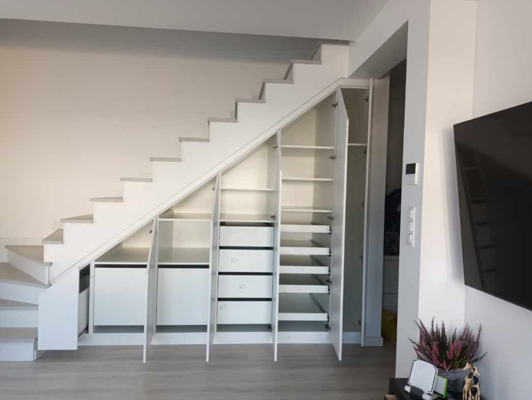 Biała szafa wnękowa pod schodami z otwartymi drzwiami i szufladami, ukazująca przestrzeń do przechowywania. Minimalistyczny design, jasne wnętrze.