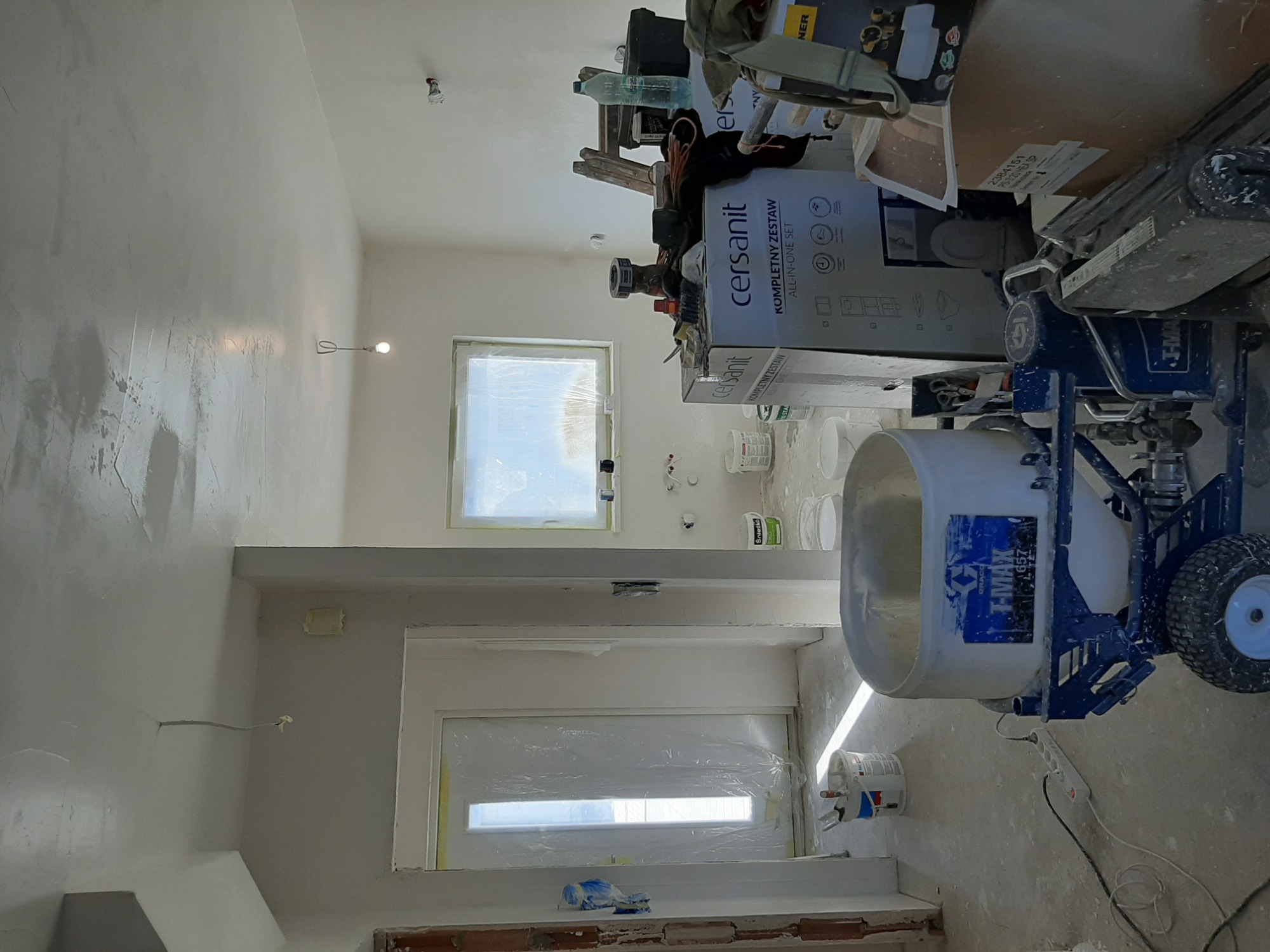 Wnętrze w trakcie prac wykończeniowych, ściana po nałożeniu gładzi, zabezpieczone okno folią malarską, widoczny agregat malarski Graco T-MAX 657 i kartony z wyposażeniem łazienki Cersanit.