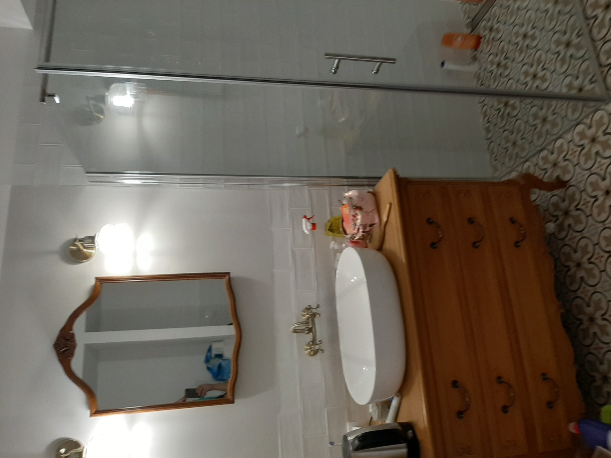 Nowoczesna łazienka z owalną umywalką nablatową, drewnianą komodą z ozdobnymi uchwytami, lustrem w drewnianej ramie i kabiną prysznicową oddzieloną szklaną taflą.