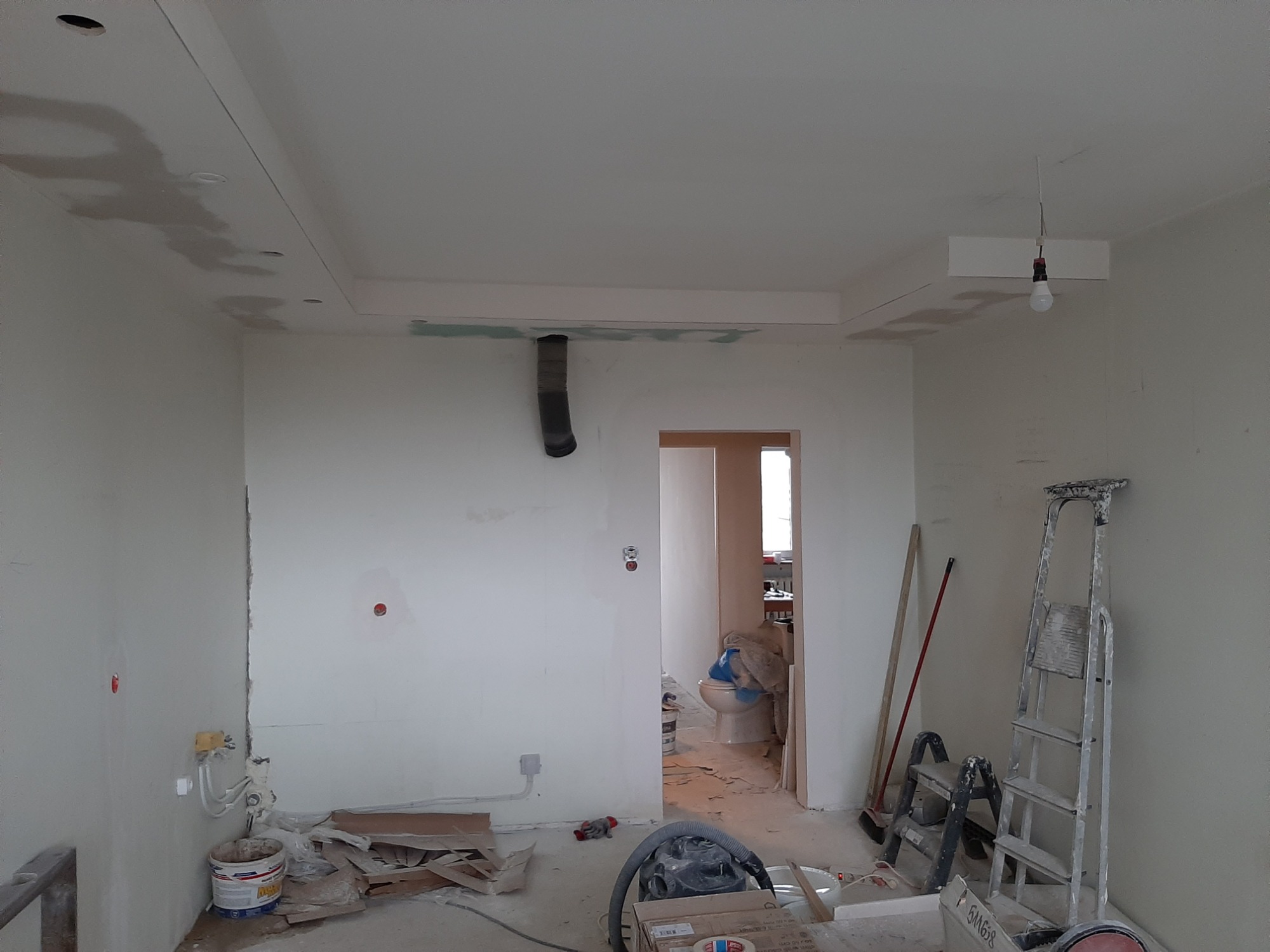 Wnętrze w trakcie remontu: ściany przygotowane do malowania, widoczne fragmenty szpachli na suficie, podwieszany sufit z otworami na oświetlenie, rura wentylacyjna na ścianie, drabina oparta...