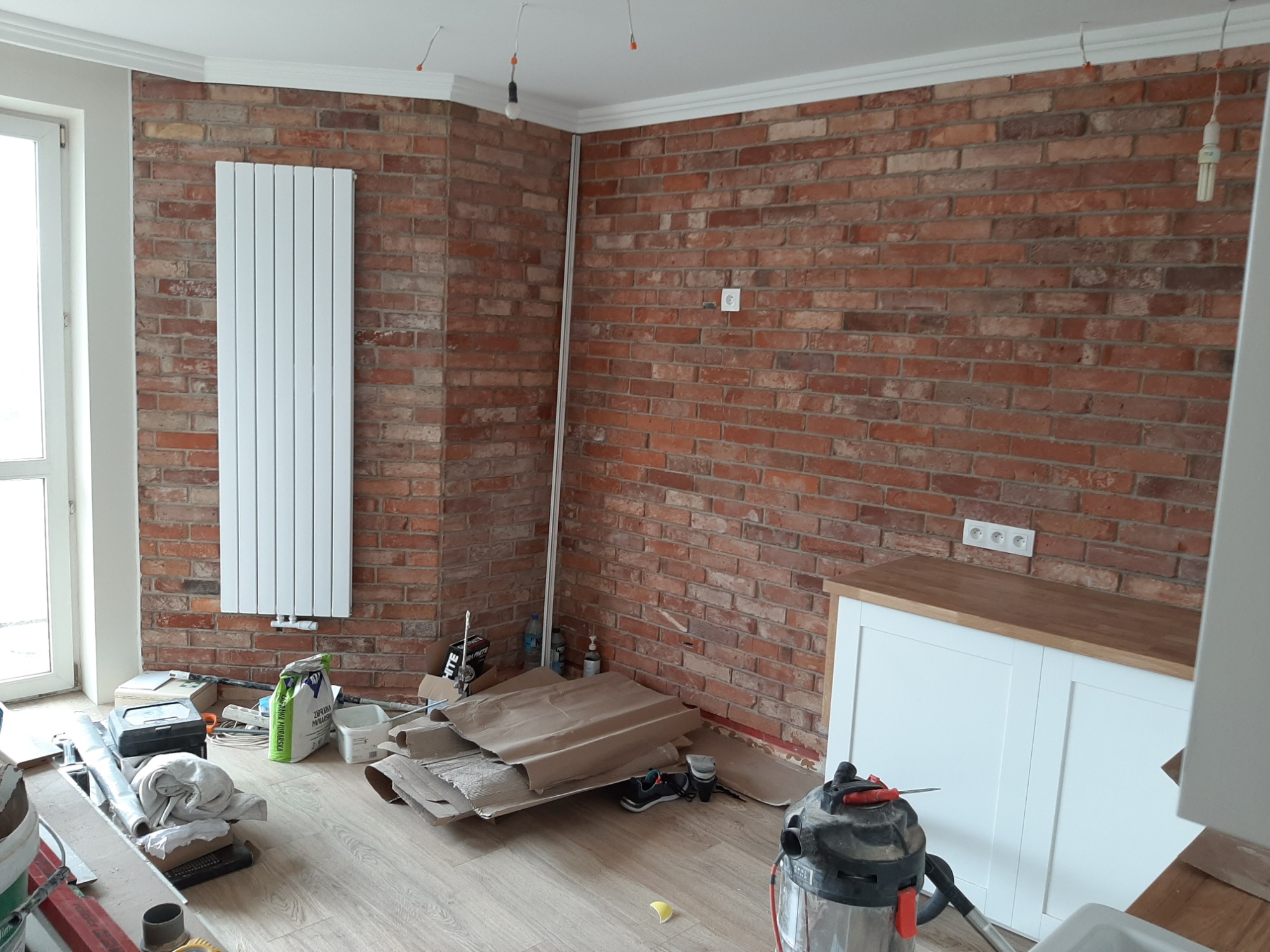 Wnętrze w trakcie remontu: ściana z czerwonej cegły z białym grzejnikiem panelowym, biała listwa przysufitowa, kartony i narzędzia budowlane na podłodze, widoczne gniazdka elektryczne.