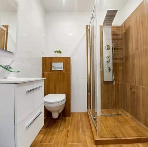 Nowoczesna łazienka z drewnopodobnymi płytkami na podłodze i ścianach kabiny prysznicowej, biała umywalka z szklaną misą i toaleta podwieszana z zieloną roślinką na górze.