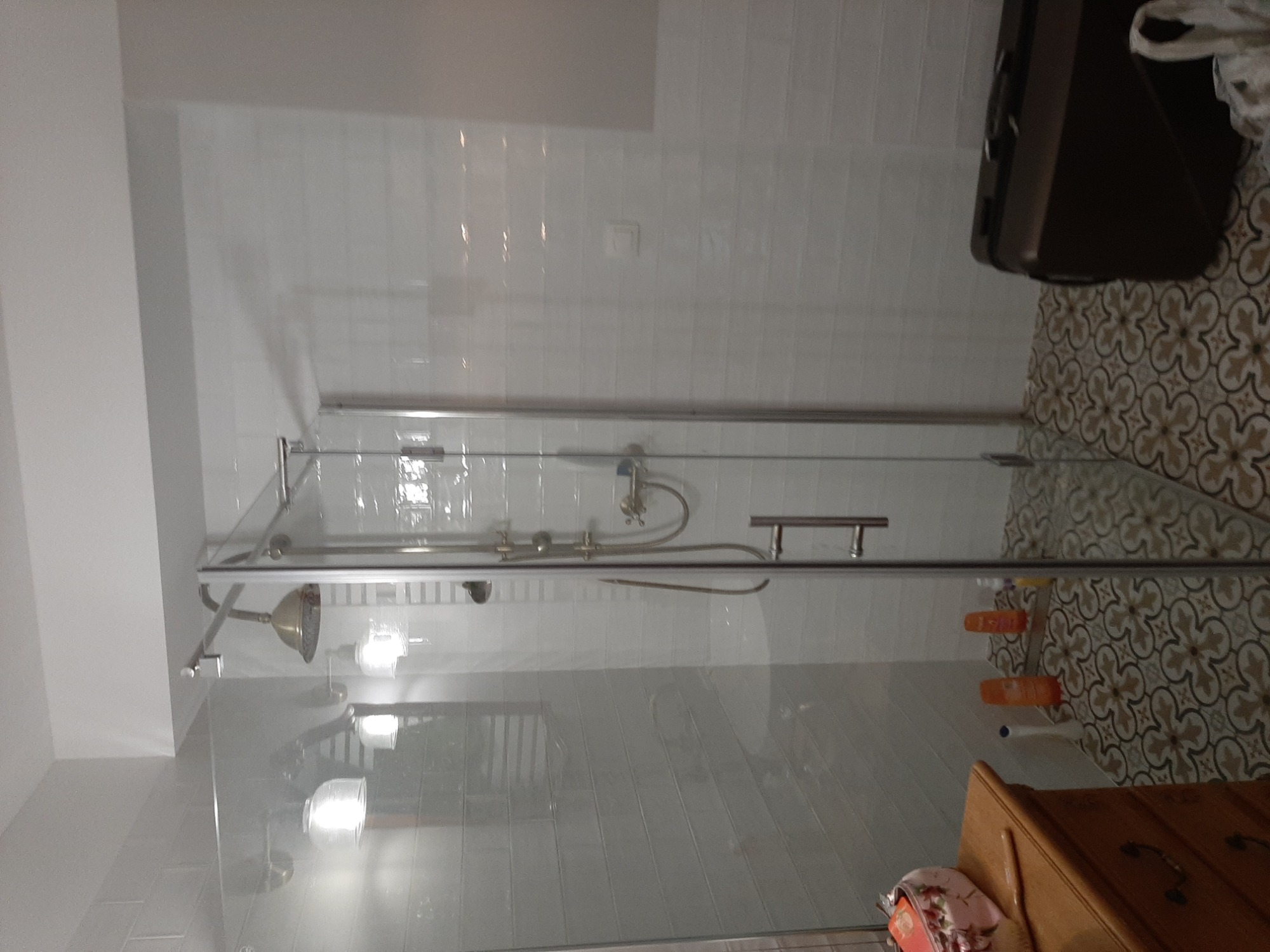 Szklana kabina prysznicowa z chromowaną armaturą na tle białych płytek i dekoracyjnej mozaiki podłogowej, widok na drewnianą komodę i torbę podróżną w rogu.