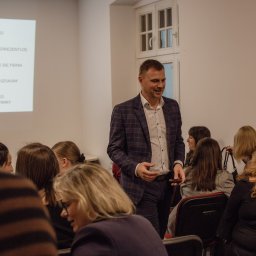 RM Business Group - Uśmiechnięty mężczyzna w kraciastej marynarce przechodzi między uczestnikami biznesowego spotkania, na ścianie widoczna projekcja z punktami do autoprezentacji.