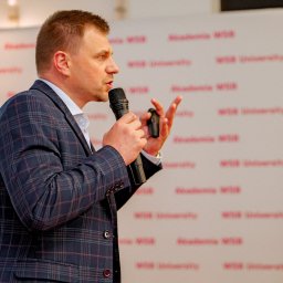 RM Business Group - Mężczyzna w granatowej marynarce w kratę, trzymający mikrofon i gestykulujący lewą dłonią, przemawia na tle białej ściany z powtarzającym się czerwonym napisem 'Akademia MIB University'.