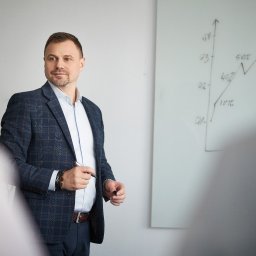 RM Business Group - Mężczyzna w eleganckiej marynarce w kratę i jasnej koszuli, z markerem w dłoni, prezentuje wykres wzrostu na białej tablicy, patrząc w stronę rozmytych słuchaczy.