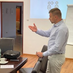 RM Business Group - Mężczyzna w koszuli w paski gestykuluje, prowadząc szkolenie z prezentacją 'Klient „Inni”' i logo RM Business Group na ekranie. Na stole projektor.