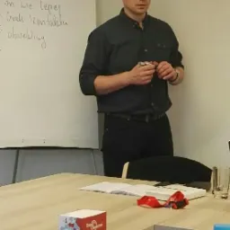 Mężczyzna w ciemnej koszuli stoi przed tablicą z notatkami w sali konferencyjnej, trzymając coś w dłoniach, na stole widoczny notatnik, pudełko i butelka wody.