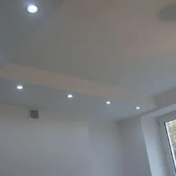 Wnętrze pokoju po malowaniu, z gładkimi białymi ścianami i sufitem, oświetlenie punktowe w suficie podwieszanym, widoczne okno z widokiem na budynek na zewnątrz, wisząca żarówka na kablu.